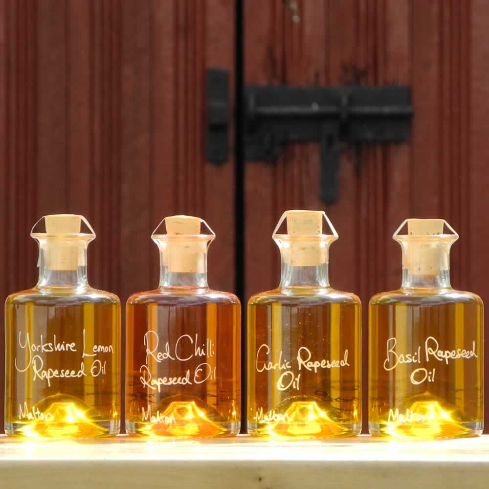 The Infused Oil Collection with Lemon Rapeseed Oil, Red Chilli Rapeseed Oil, Garlic Rapeseed Oil and Basil Rapeseed Oil in refillable 200ml bottles, cooking gift set displayed in front of red barn doors